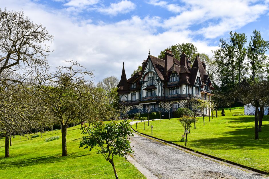 A large Victorian-style house with decorative timber framing, dark roofing, and multiple gables and chimneys, surrounded by a well-maintained garden. The garden features a neatly trimmed lawn with lus