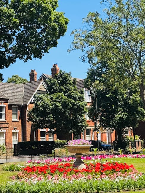 A well-maintained urban garden scene featuring a vibrant flower bed with red, pink, and white blooms in the foreground, surrounded by lush green grass. In the middle ground, there is a decorative ston